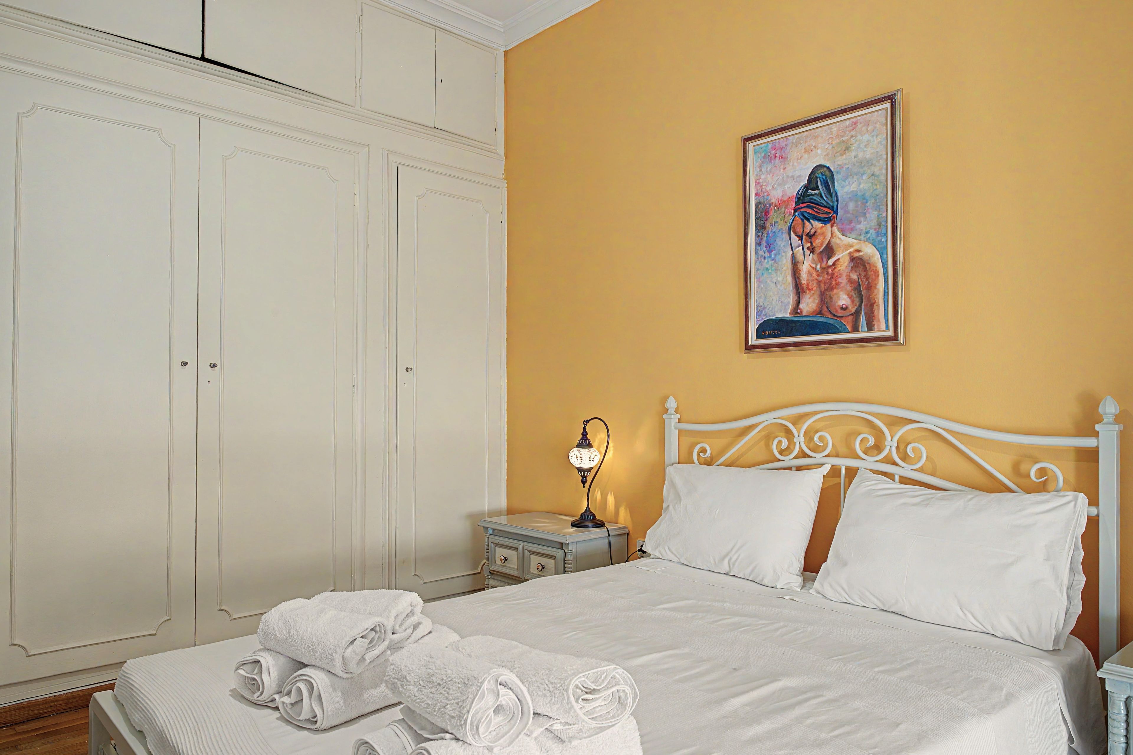 A bedroom with a bed, white curtains, a nightstand with a lamp, and a painting on the wall.
