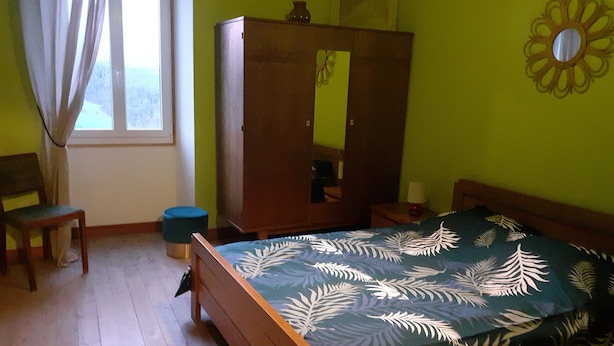 A bedroom with a bed, a wooden wardrobe, a chair, a round stool, and a window with curtains.