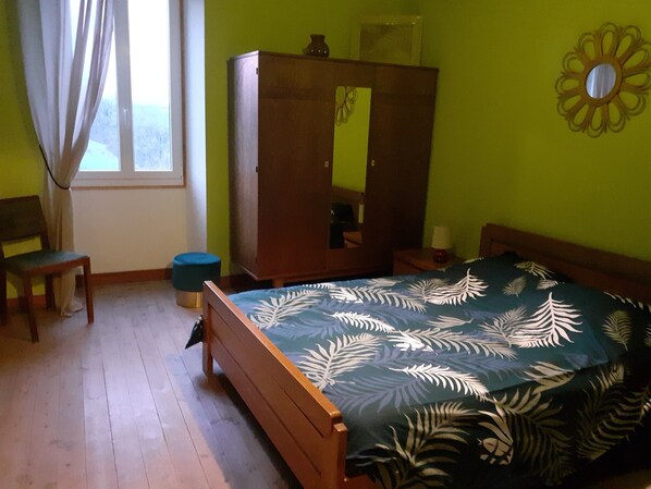 A bedroom with a bed, a wooden wardrobe, a chair, a round stool, and a window with curtains.
