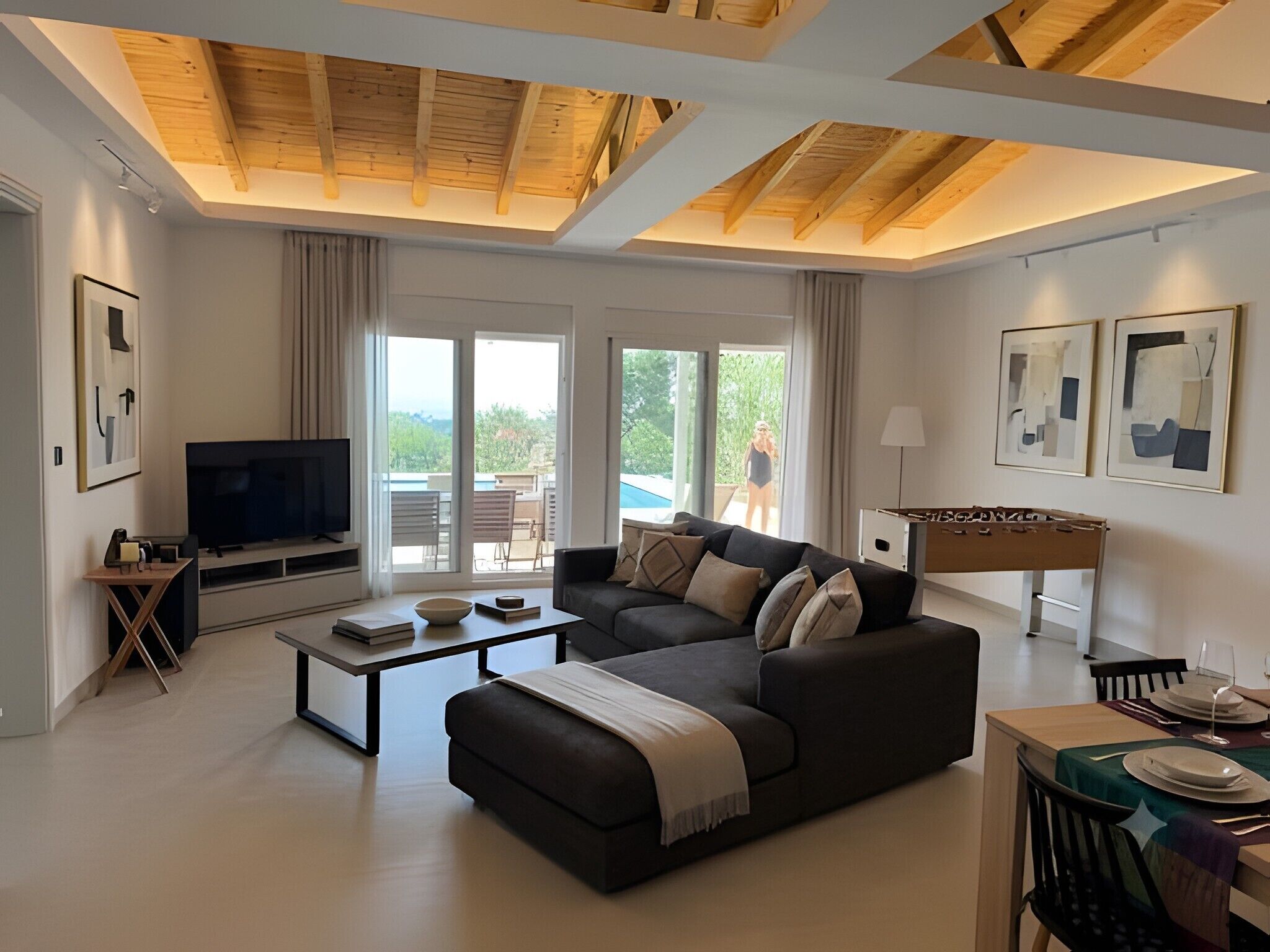 A modern living room with a sofa, coffee table, and a television. There is a balcony with a view of greenery.