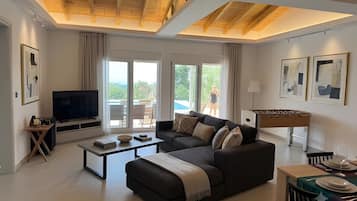 A modern living room with a sofa, coffee table, and a television. There is a balcony with a view of greenery.