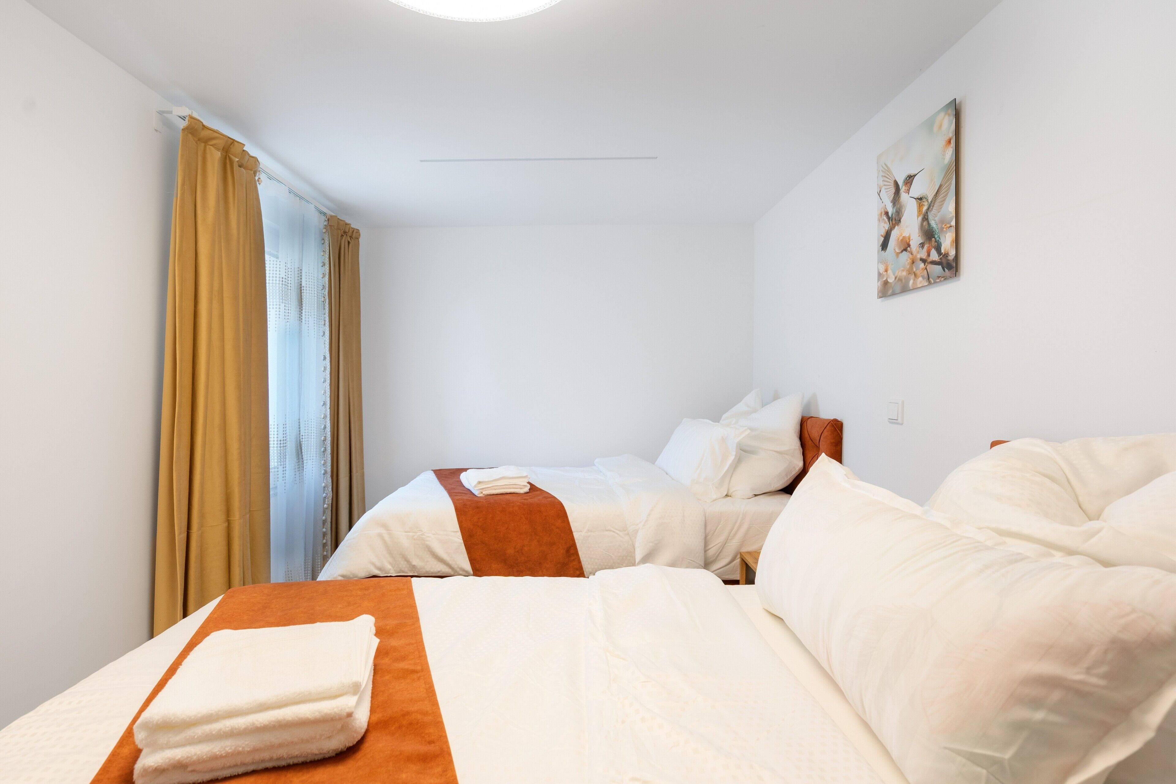 Ein Hotelzimmer mit zwei Betten, einem Fenster mit Vorhängen, einem Gemälde an der Wand und einem Lichtschalter.