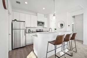 A modern kitchen with white cabinetry, stainless steel appliances, and a central island with bar stools. - New Build: B: 1BR/1BA Modern Condo in Houston (Houston)