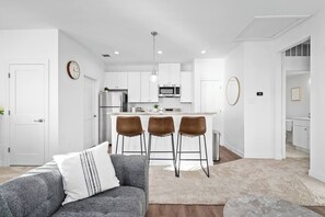Eine moderne Küche mit weißen Schränken, einer Mikrowelle und einem Edelstahl-Kühlschrank. Drei braune Barhocker und eine weiße Arbeitsplatte sind vorhanden.
