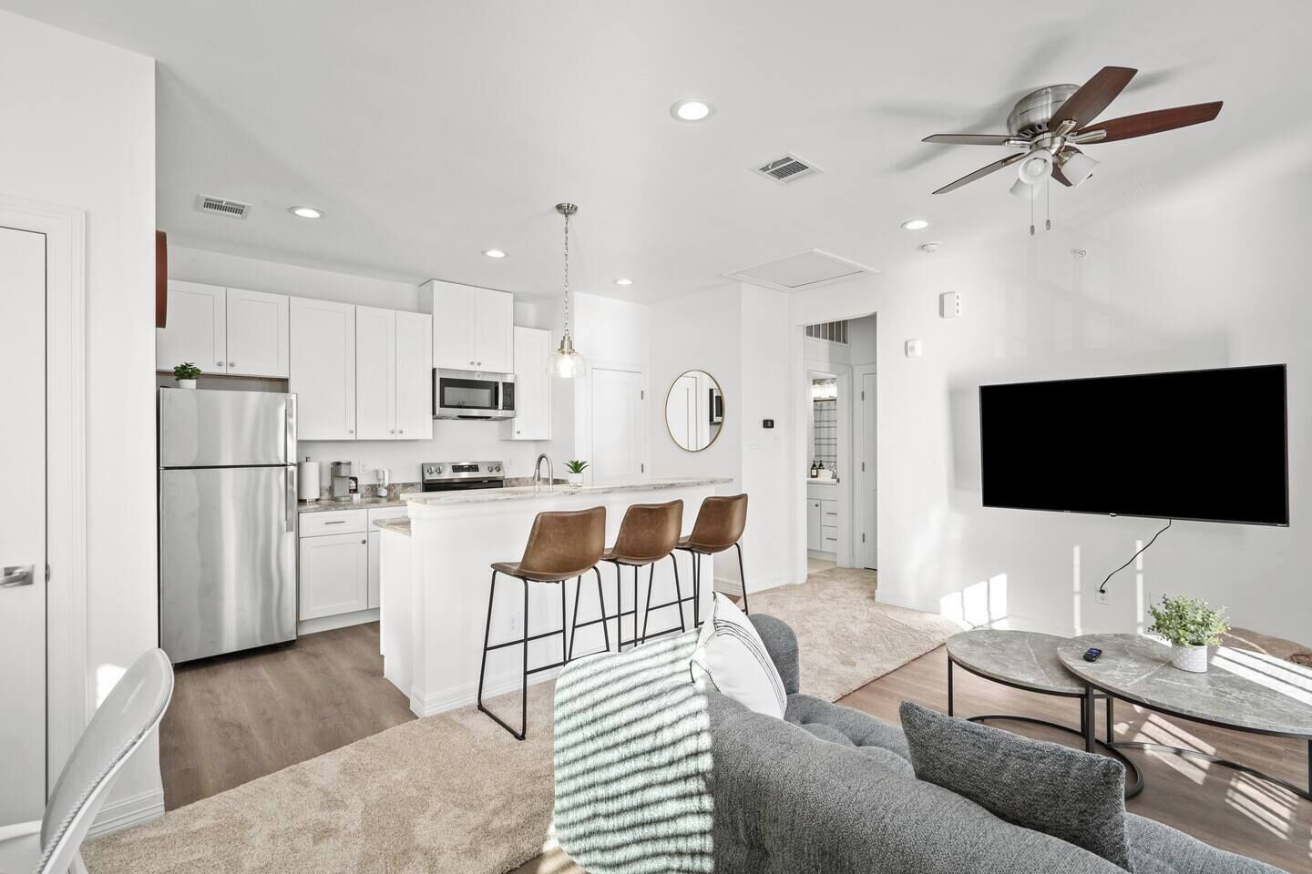 A modern kitchen with white cabinets, a refrigerator, and a microwave. There is a dining area with a bar stool and a television mounted on the wall.