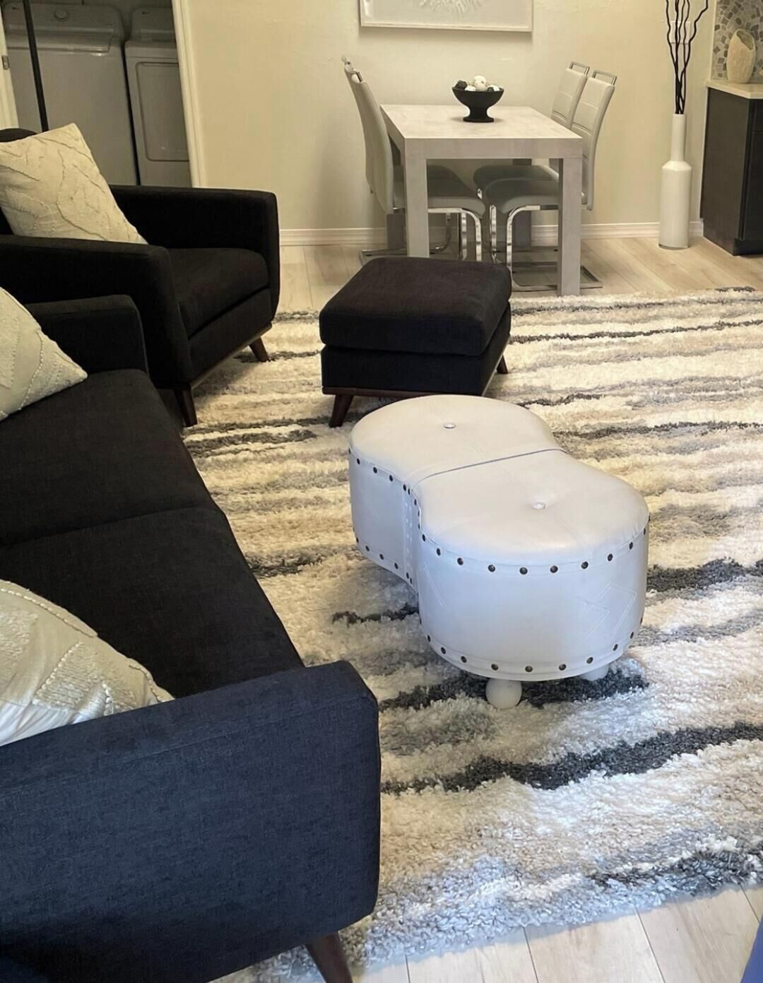 A living room with a black sofa, a white ottoman, a striped rug, and a dining area with a table and chairs.
