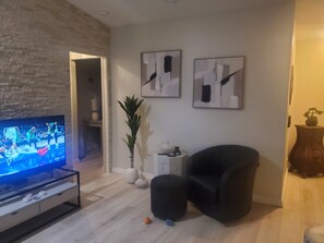 A living room with a flat-screen TV, a black armchair, a potted plant, and abstract wall art. - Gorgeous modern 2 bd, 2 full bth condo within walking distance of Cape Harbour! (Cape Coral)