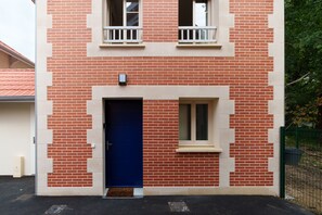 Un edificio de dos pisos con fachada de ladrillo rojo, una puerta azul y una ventana pequeña con marco blanco.