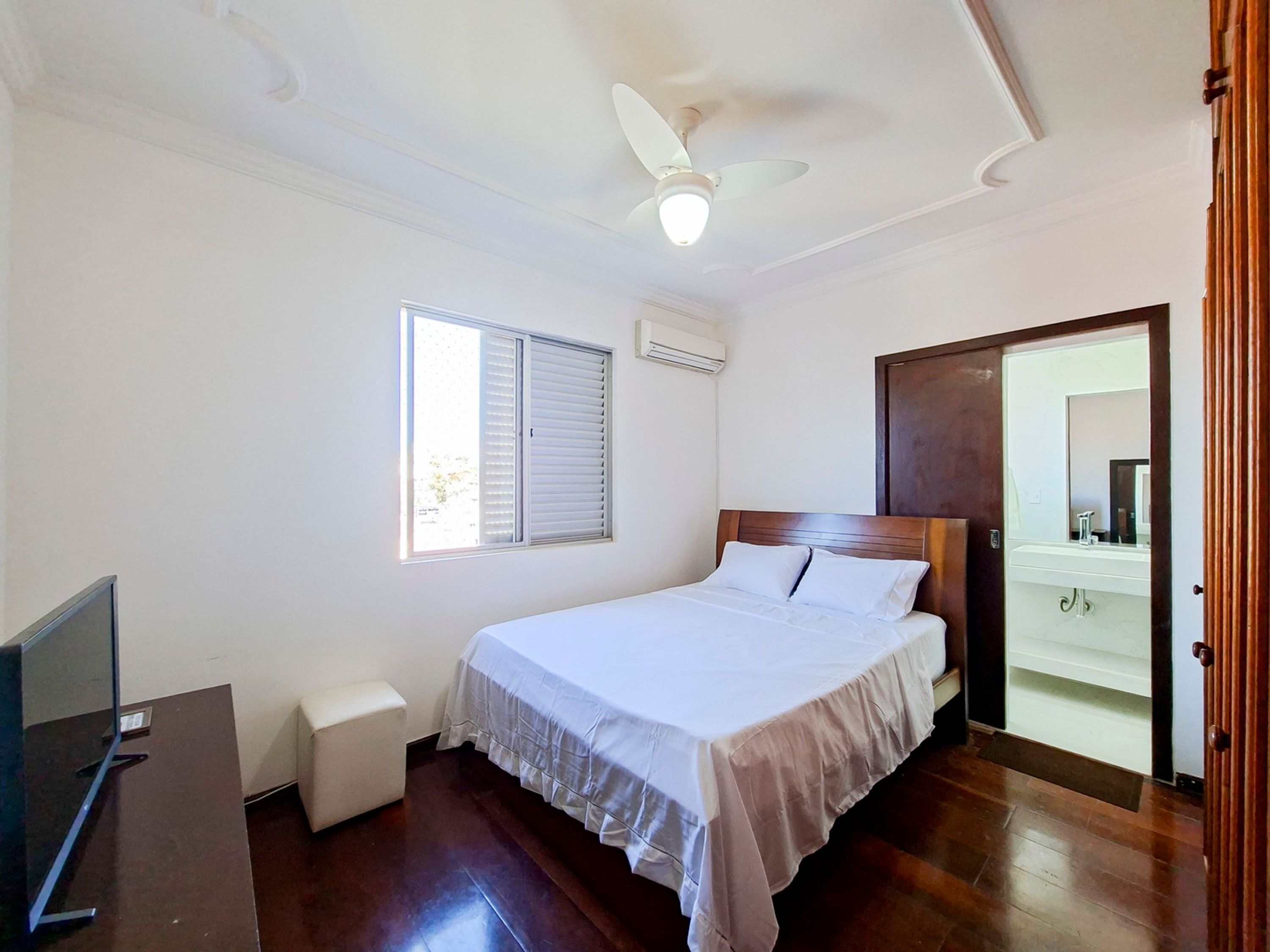 A bedroom with a bed, a ceiling fan, an air conditioning unit, a window with blinds, a wooden door, and a small bedside table.