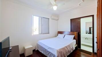 A bedroom with a bed, a ceiling fan, an air conditioning unit, a window with blinds, a wooden door, and a small bedside table.