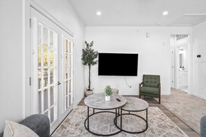Una sala de estar con televisor de pantalla plana, dos mesas redondas, un sillón verde y una planta en maceta.