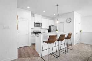 Cocina moderna con armarios blancos, una isla central con taburetes, microondas, estufa y refrigerador.