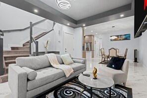 Ein modernes Wohnzimmer mit einem grauen Sofa, einem runden Couchtisch und einem Essbereich mit einem weißen Tisch und Stühlen.