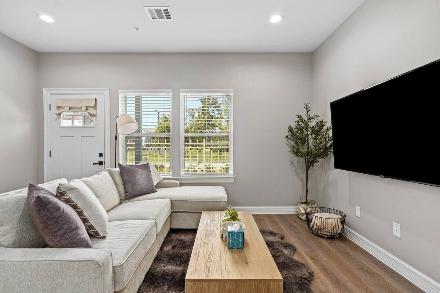 A living room with a sectional sofa, a coffee table, a TV, and a large window with blinds.