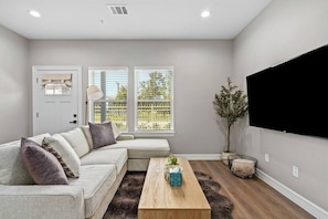 A living room with a sectional sofa, a coffee table, a TV, and a large window with blinds. - New Build 2-Story Townhome/Single Master Suite (Houston)
