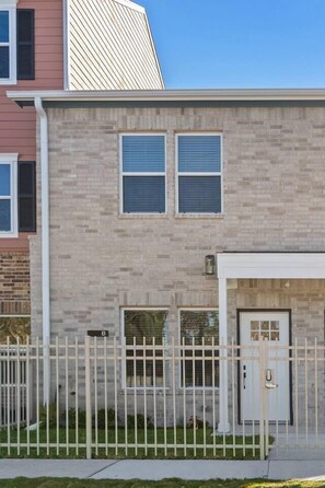 A two-story residential building with a brick facade, a white door, and a small front yard with a white fence. - New Build 2-Story Townhome/Single Master Suite (Houston)