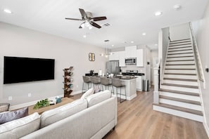 A modern kitchen with a central island, a dining area with bar stools, a living room with a sofa, and a staircase. - New Build 2-Story Townhome/Single Master Suite (Houston)