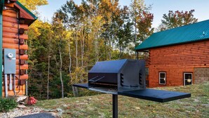 Puinen mökki, jossa on metallikatto, ulkona oleva grilli ja sen vieressä pöytä sekä taustalla metsä.