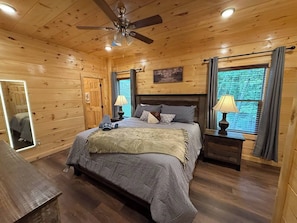 Ein Schlafzimmer einer Holzhütte mit einem Bett, zwei Nachttischen, einem Deckenventilator und einem Fenster mit Vorhängen.