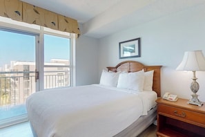 A hotel room with a bed, a bedside table with a lamp, a framed picture on the wall, and a large window with a view of buildings. - Carolinian Beach Resort 1833 (Myrtle Beach)