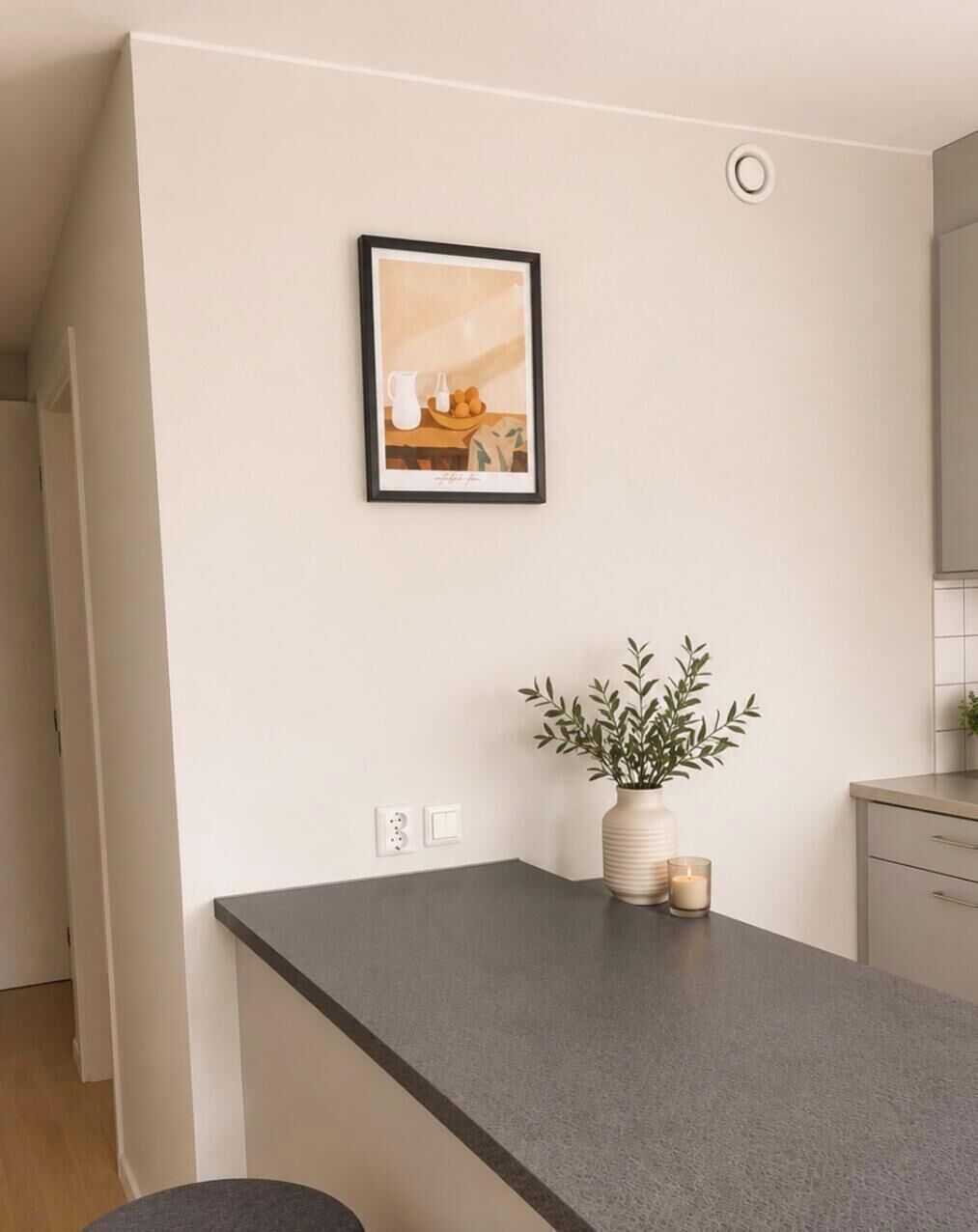 Une cuisine avec un plan de travail gris, un tableau encadré au mur, une plante en pot et une bougie allumée.