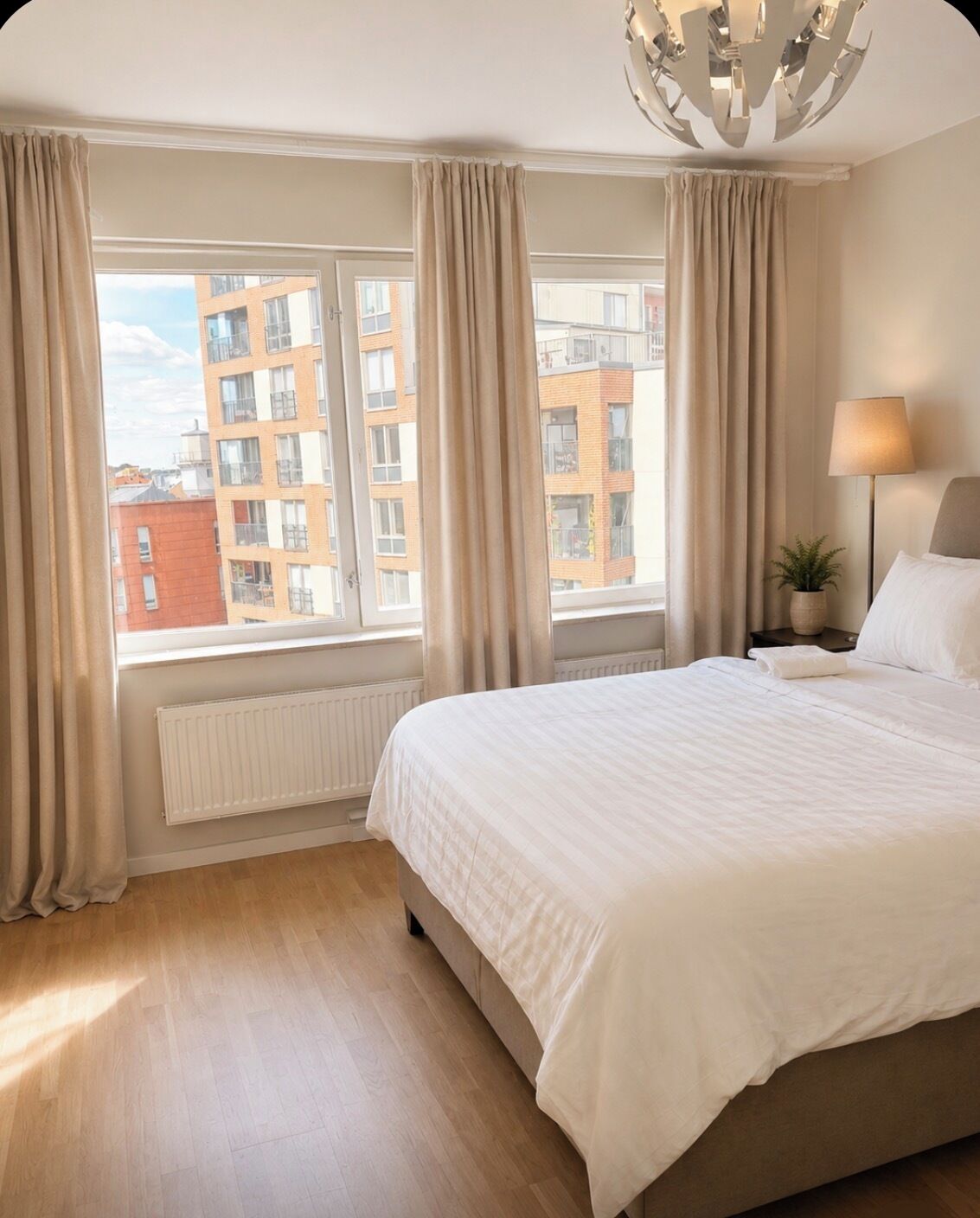 Et hotelværelse med en stor seng, en radiator, et vindue med gardiner, en lampe og udsigt til bygninger.