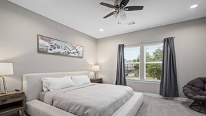 A bedroom with a bed, a nightstand, a lamp, a ceiling fan, a window with curtains, and a chair. - Newly Built 2-Story Townhome: Single Master Suite (Houston)