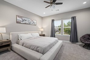 Ein Schlafzimmer mit Bett, Nachttisch, Lampe, Deckenventilator, Fenster mit Vorhängen und einem Sessel.