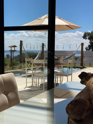 Vista desde una habitación con un ventanal grande que da a un área de piscina con mobiliario exterior y una hamaca.