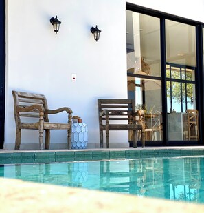 A poolside area with two wooden chairs, a hexagonal planter, and a glass door leading to an interior space.