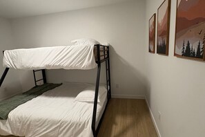A bunk bed with a white mattress and a green blanket, a wooden floor, and three framed pictures on the wall. - New 3BR Lake Country Home | Golf, Wine, Lakes & Mountain Getaway (Kelowna)