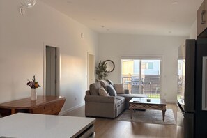A modern living room with a sectional sofa, a dining table, and a kitchen area with a refrigerator. - New 3BR Lake Country Home | Golf, Wine, Lakes & Mountain Getaway (Kelowna)