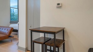 Una habitación con un sofá de cuero marrón, una mesa de madera con estantes, un termostato en la pared y una ventana con vistas a edificios y árboles.