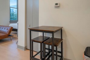 A room with a brown leather sofa, a wooden table with shelves, a thermostat on the wall, and a window with a view of buildings and trees. - Modern Studio - Nearby Parking - Downtown - 3F (Louisville)