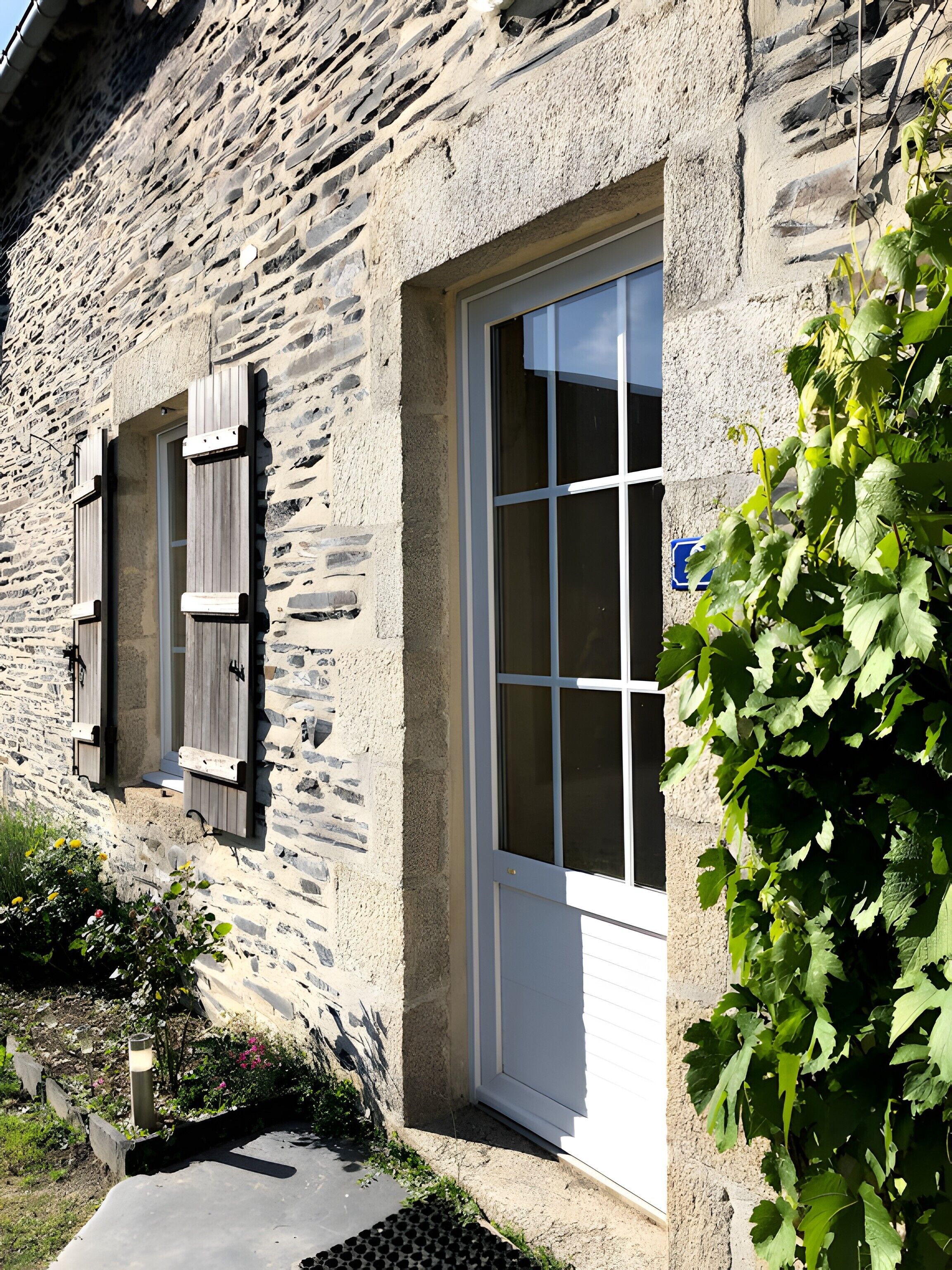 Ein Steinhaus mit weißer Tür und Fensterläden, ein Fenster und ein Garten mit Pflanzen und Blumen.