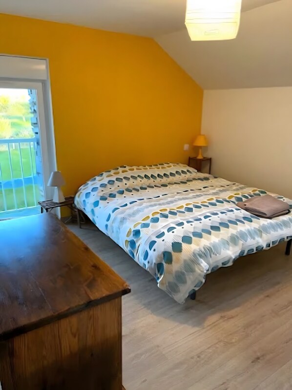A bedroom with a bed, a wooden desk, a lamp, and a window with a view of greenery.