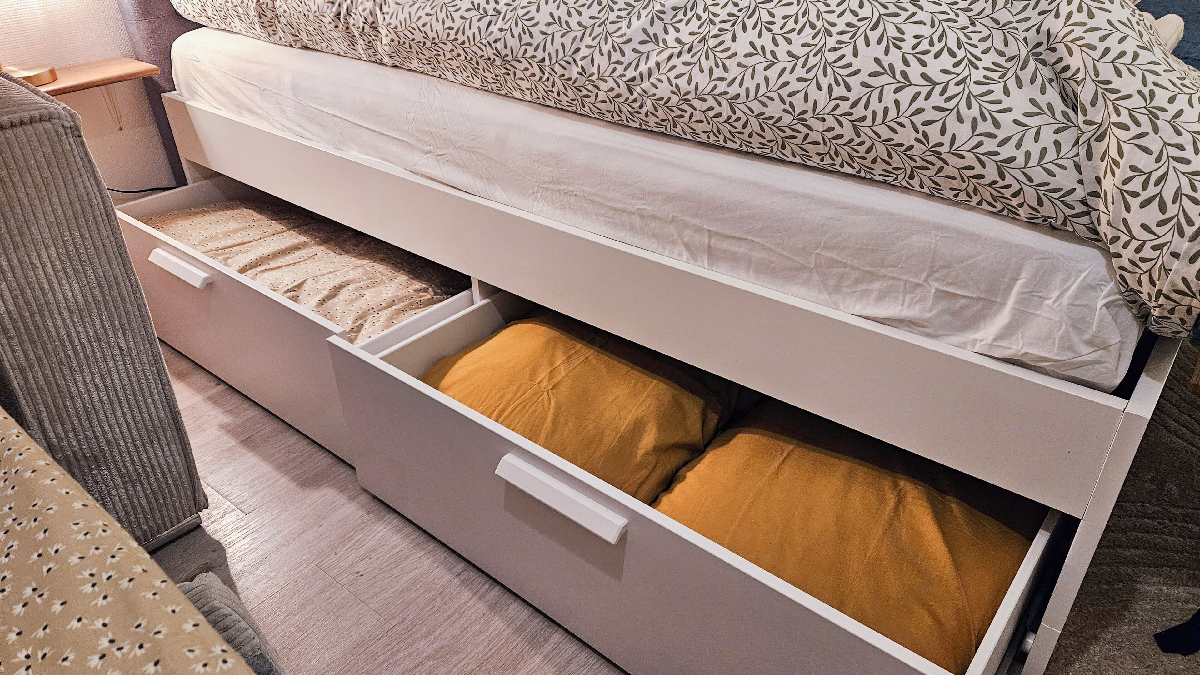 Un lit avec une tête de lit à motifs, un matelas blanc et un tiroir de rangement avec un coussin marron.