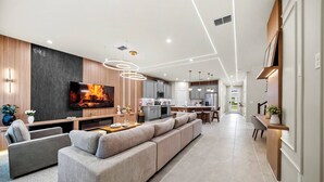 Een moderne woonkamer met een grote flatscreen televisie, een bank en een eethoek, met een keuken op de achtergrond.
