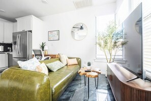 A modern living space with a green sofa, a wooden coffee table, a kitchen area with a refrigerator, and a large window with blinds. - Gated 2BR | Modern Austin Home | Near DT | EV Plug (Austin)