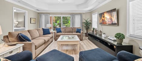 A living room with a beige sofa, a coffee table, and two armchairs. There is a ceiling fan, a TV mounted on the wall, and a window with curtains.