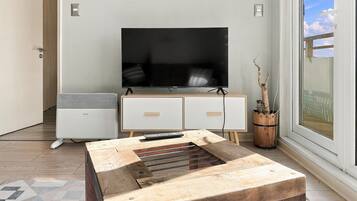 A modern living room with a flat-screen TV, a small white cabinet, a wooden coffee table, and a large window with a view.