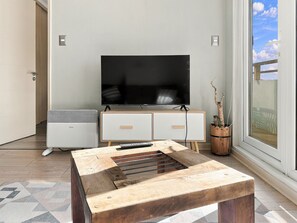 Una sala de estar moderna con un televisor de pantalla plana, un mueble blanco pequeño, una mesa de centro de madera y un ventanal con vistas.