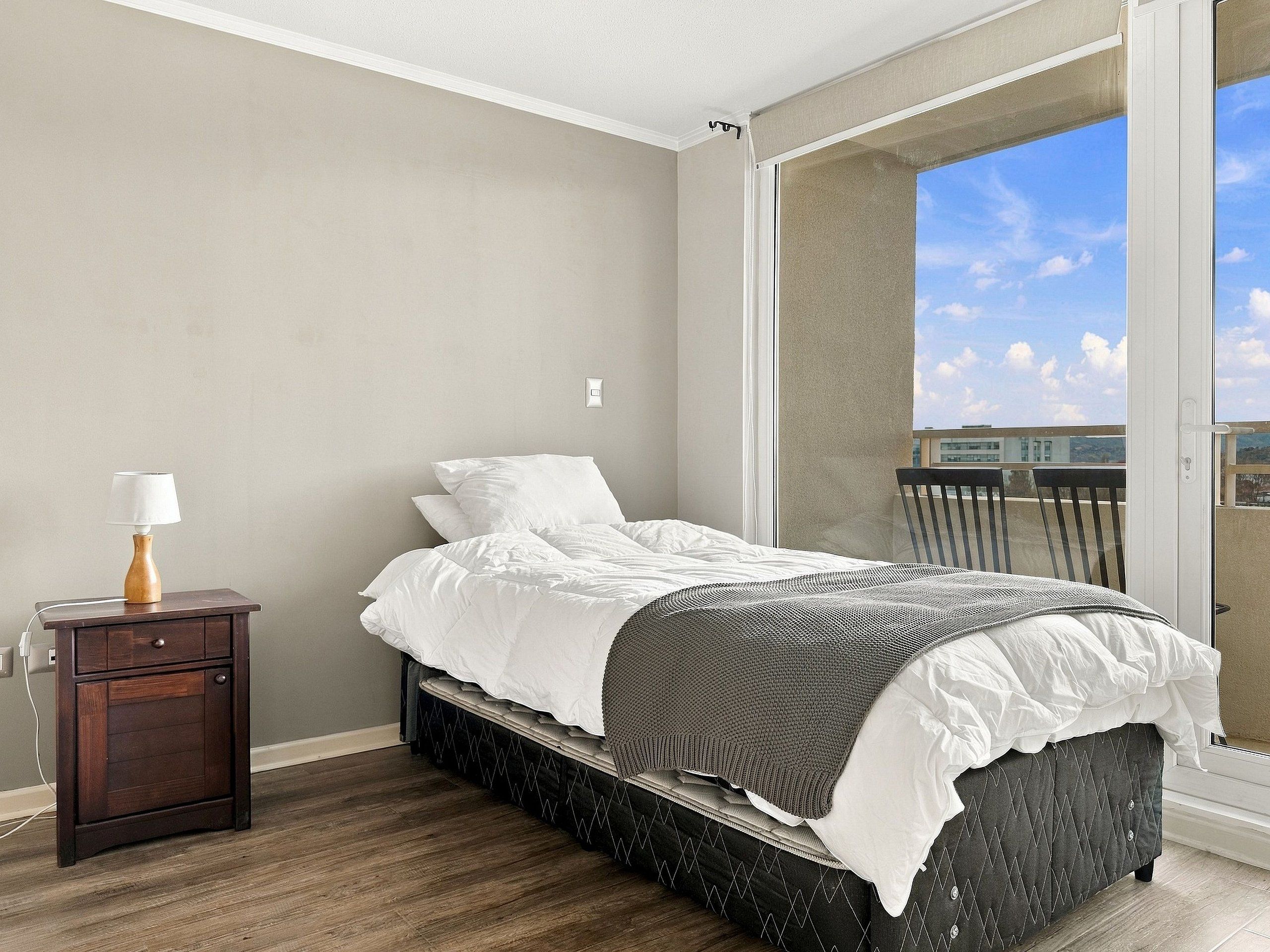 A bedroom with a bed, a nightstand, a lamp, and a view of the outside.