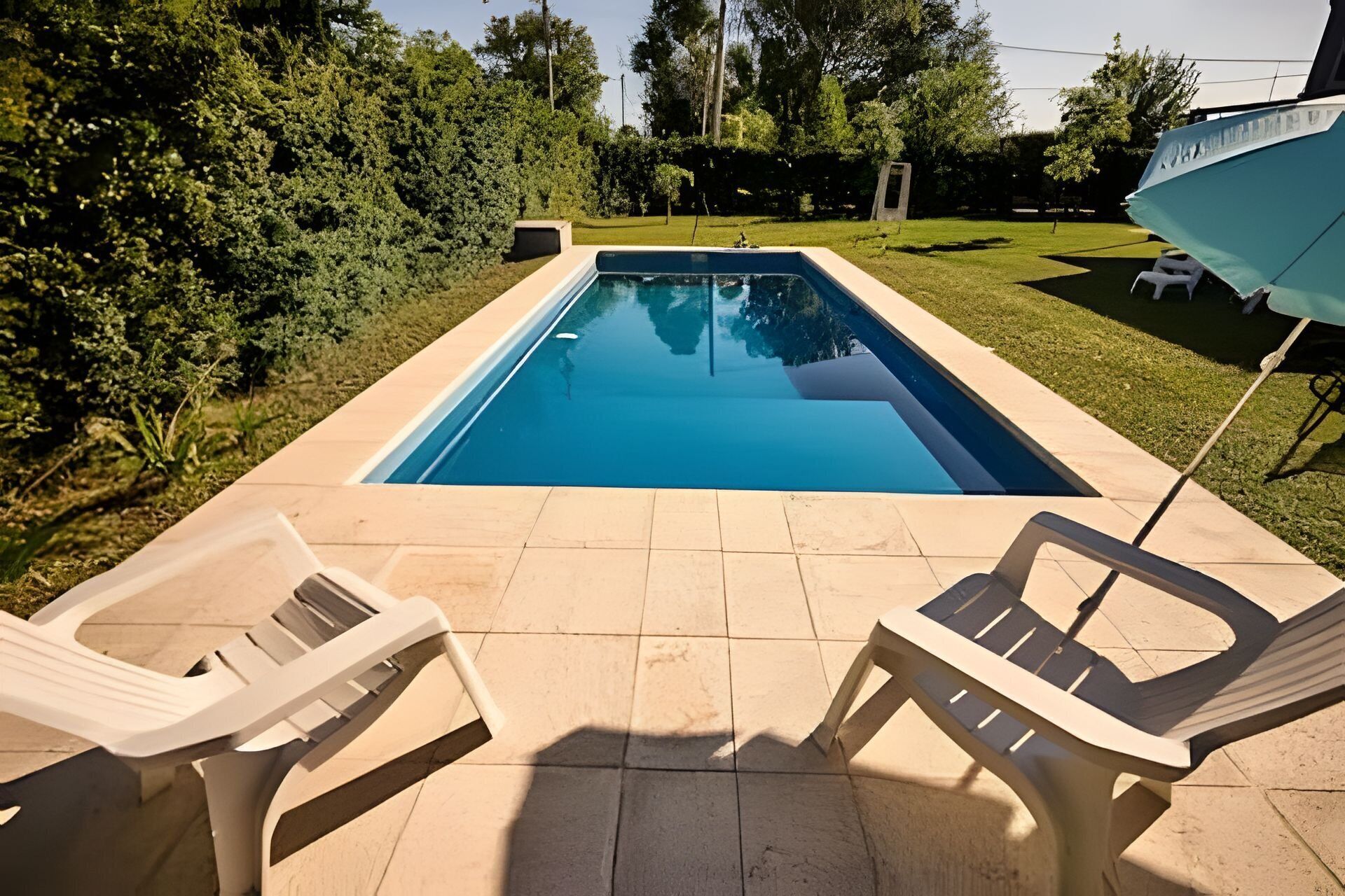 Una piscina rectangular con solárium empedrado, rodeada de follaje y dos sillas de descanso blancas.