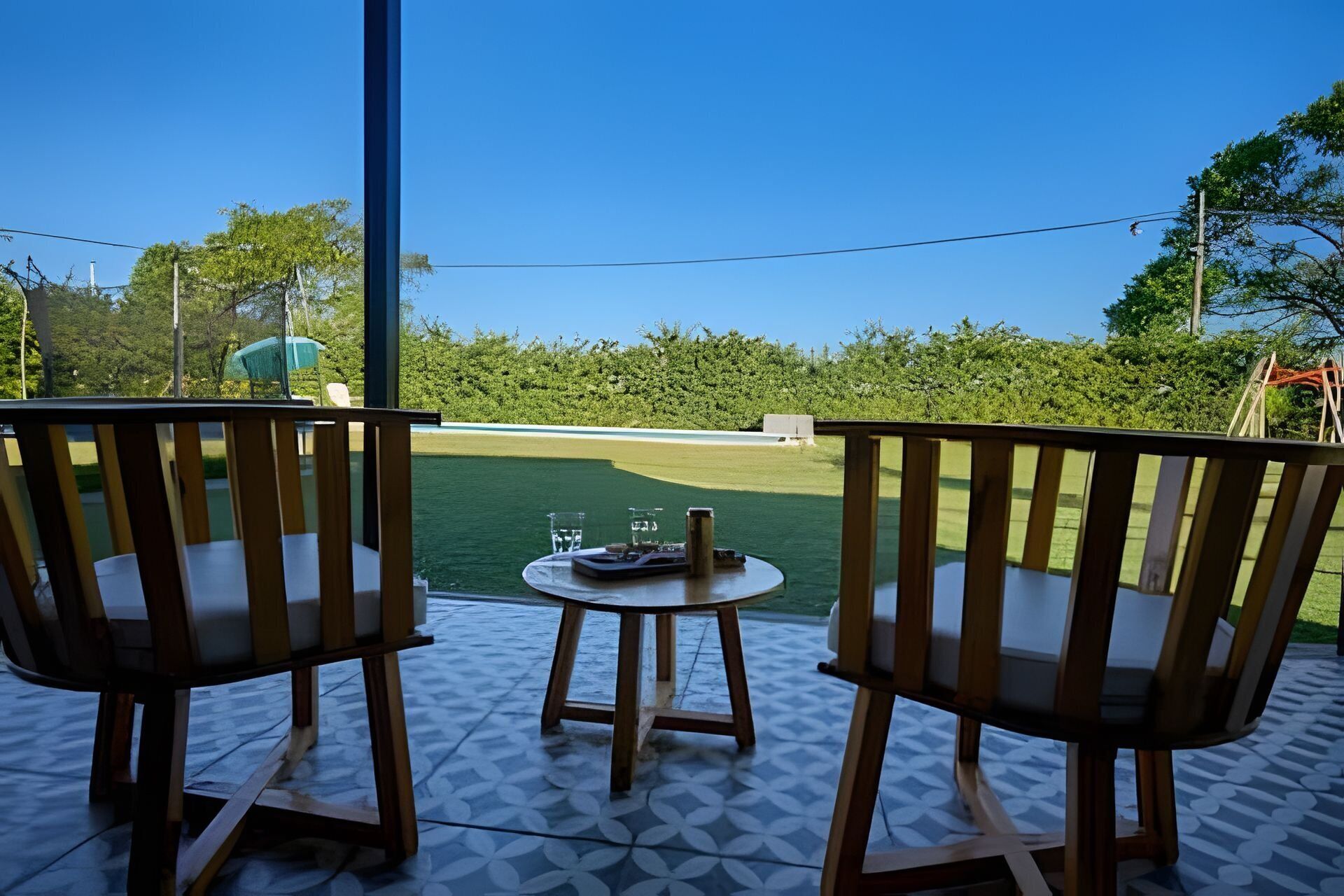 Two wooden chairs facing a small table with a glass and a bottle, overlooking a green lawn and trees.