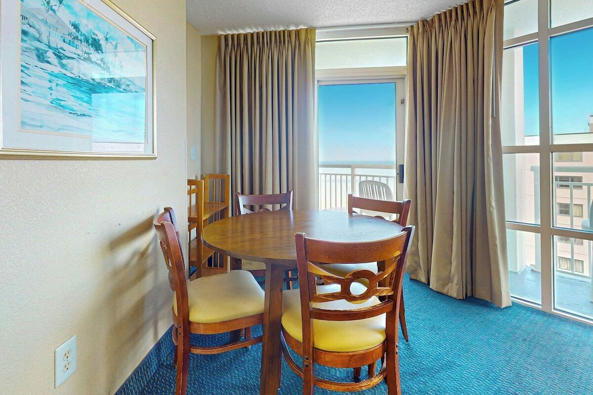 A hotel room with a round table and chairs, a large window with curtains, and a framed picture on the wall.