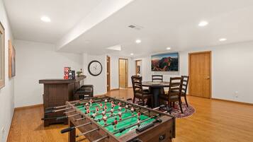 A room with a foosball table, a dining table with chairs, a clock on the wall, and a painting.