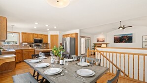 A modern kitchen with wooden cabinets, a dining area set for six with a wooden table, and a view of a mountain landscape through a window.