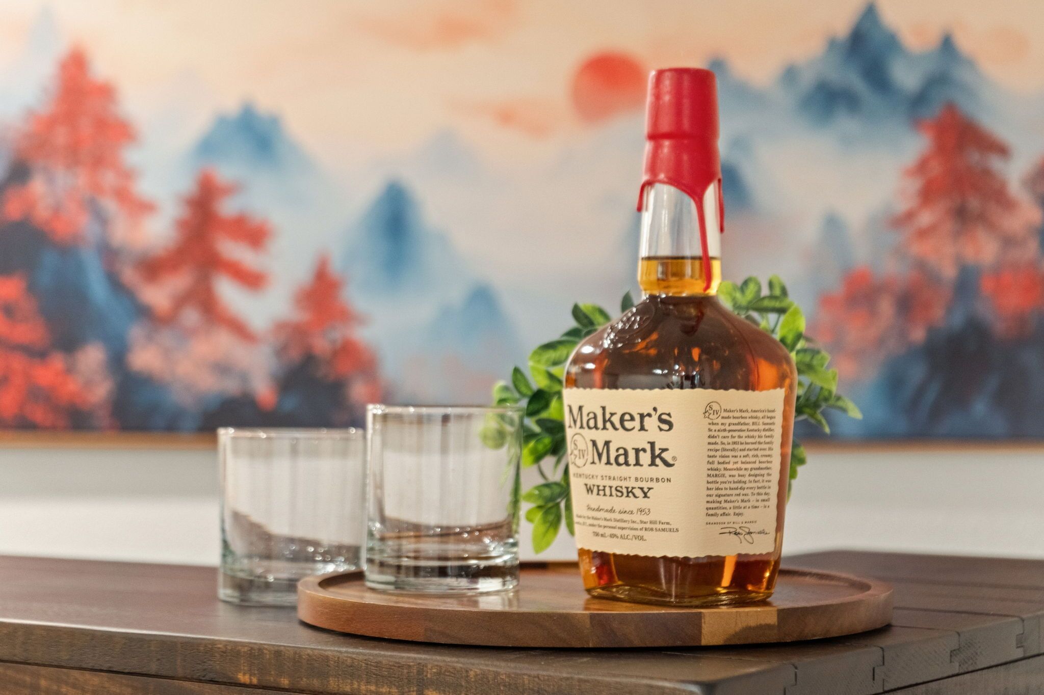 A bottle of Maker's Mark bourbon whiskey on a wooden coaster with two glasses beside it.