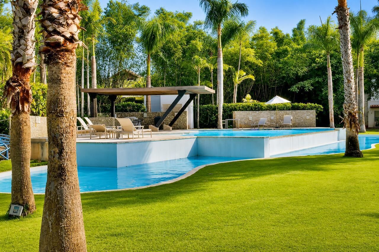 Una piscina con zona de descanso, rodeada de palmeras y un césped cuidado.